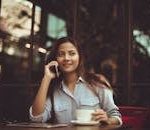 Young woman in casual shirt talking on phone and holding coffee at an outdoor café table.